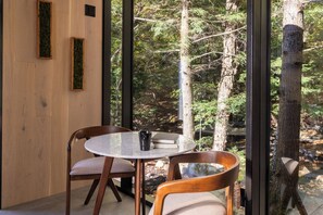 A dining area with a round table and two chairs, a wooden wall, and a view of a forest through large windows. - The Moss Rock | Mirror Cabin + Spa at Uncommon Accommodations - Smuggler's Notch (Cambridge)