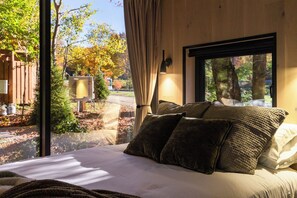 Une chambre avec une grande fenêtre, un lit recouvert de plusieurs oreillers et une vue sur des arbres et un bâtiment à l’extérieur.