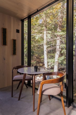 Un intérieur moderne avec une table ronde en verre, deux chaises en bois et une vue sur une forêt, visible à travers de grandes fenêtres.