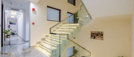 A modern interior with a marble staircase, glass railings, and a hallway leading to a door.