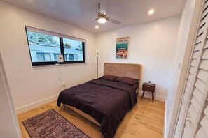 Un dormitorio con cama, ventana, ventilador de techo, mesita de noche y un cartel en la pared.