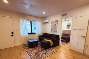 Un espacio habitable compacto con un sofá, una mesita redonda y una alfombra. Hay una ventana con cortinas, un equipo de aire acondicionado y un cartel en la pared.