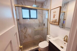 Un baño moderno con cabina de ducha de vidrio, inodoro, lavamanos con grifo dorado y espejo con un cuadro enmarcado.