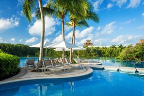 Zona junto a la piscina con sillones de descanso y sombrillas, rodeada de palmeras y un cielo despejado.