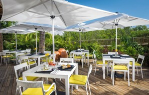 Un área de comedor al aire libre con mesas blancas y sillas amarillas, rodeada de grandes sombrillas blancas.