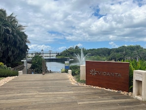 Uma passarela de madeira leva a uma placa com a palavra 'VIDANTA', uma fonte de água e uma ponte ao fundo.