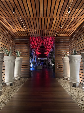 Um corredor de madeira com interior iluminado em vermelho, que leva a uma área de estar com mesas e cadeiras.