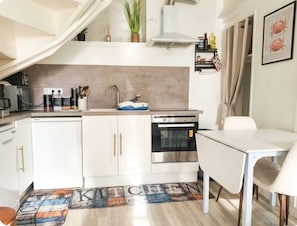 Ett kompakt kök med en inbyggd ugn, en diskho och en matplats med ett vitt bord och en stol.