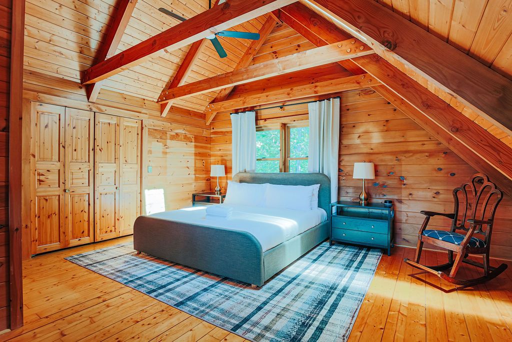 Un dormitorio de cabaña de madera con cama, mesitas de noche, una mecedora y una ventana con cortinas.