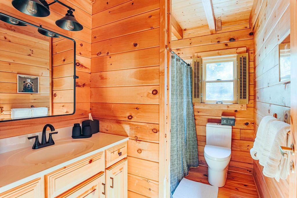 A wooden cabin bathroom with a sink, mirror, toilet, and a window with shutters.
