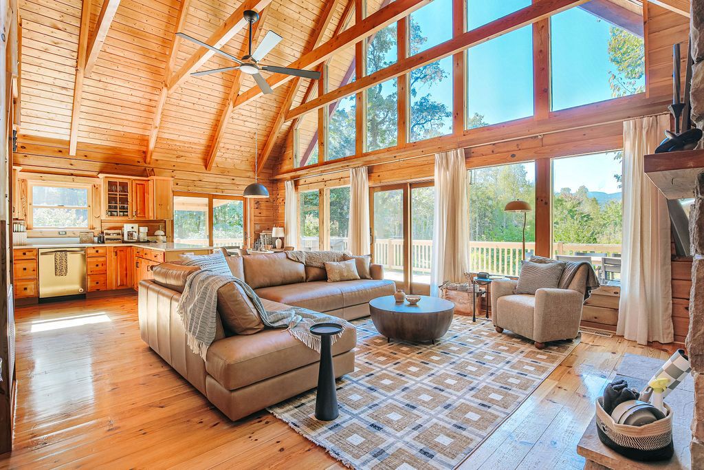 A spacious living area with a large sofa, a coffee table, and a fireplace. There is a kitchen area with wooden cabinets and a large window.
