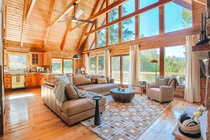 A spacious living area with a large sofa, a coffee table, and a fireplace. There is a kitchen area with wooden cabinets and a large window.