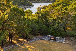A natural landscape with a stone fire pit surrounded by trees and a body of water in the background. - Panoramic Lake Views, Hot Tub, Expansive Decks - Sleeps 8, Kayaks, Dogs Welcome (Canyon Lake)