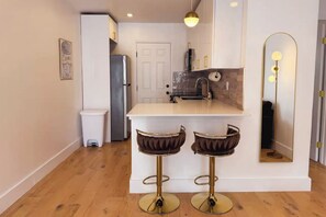 Una cocina moderna con encimera blanca, dos taburetes de bar, un refrigerador y un espejo con iluminación.