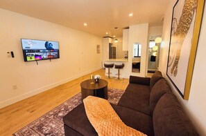 Una sala de estar con un sofá, una mesa de centro, un televisor montado en la pared y una cocina al fondo.