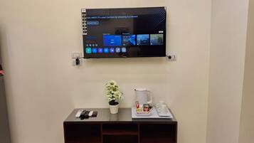 A wall-mounted TV displaying a user interface with various options. Below the TV, there is a small table with a potted plant, a coffee maker, and a tray with cups and condiments.