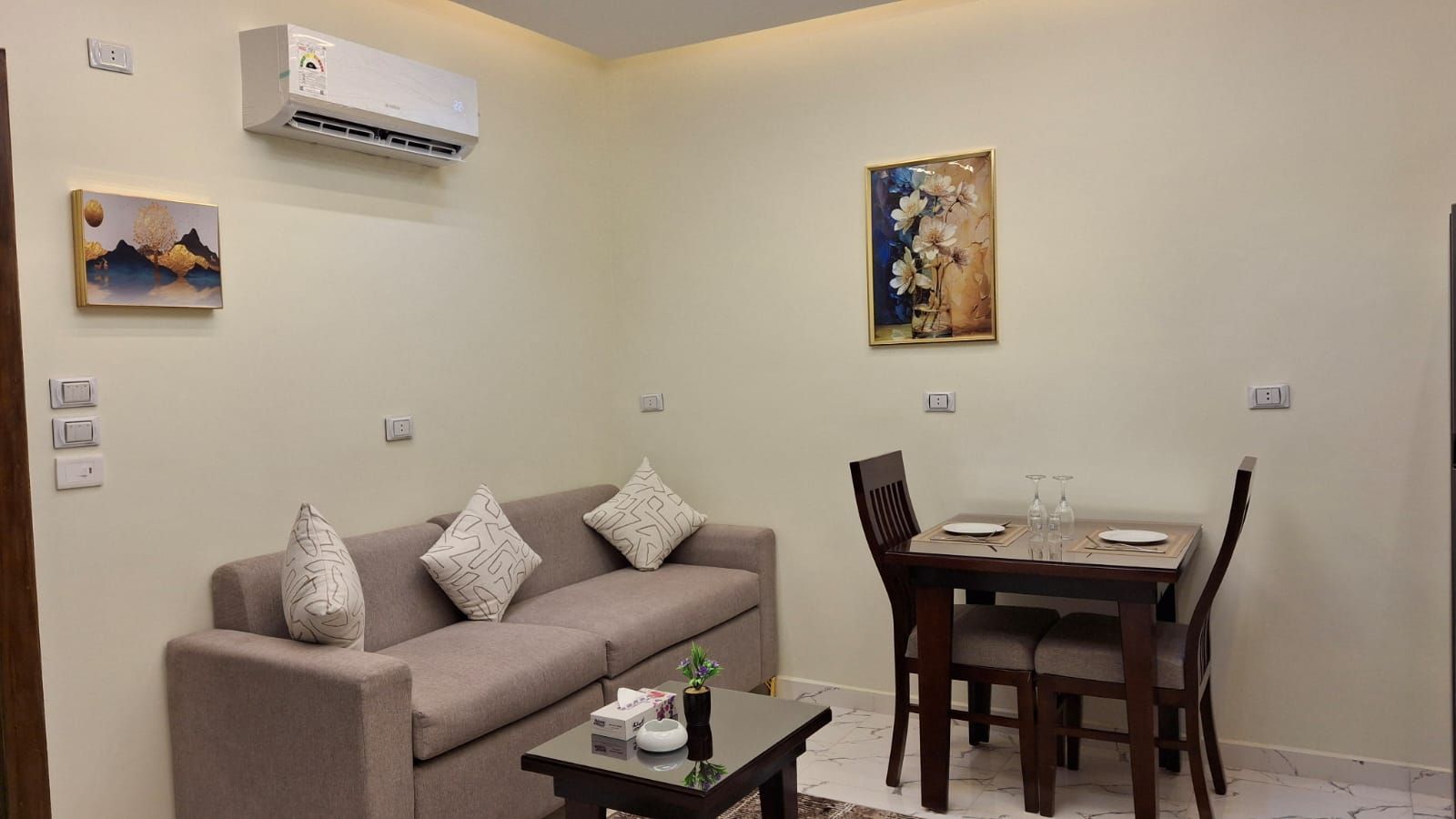 Een woonkamer met een bank, een salontafel en een eettafel met stoelen. Er hangt een airconditioner aan de muur en twee schilderijen.
