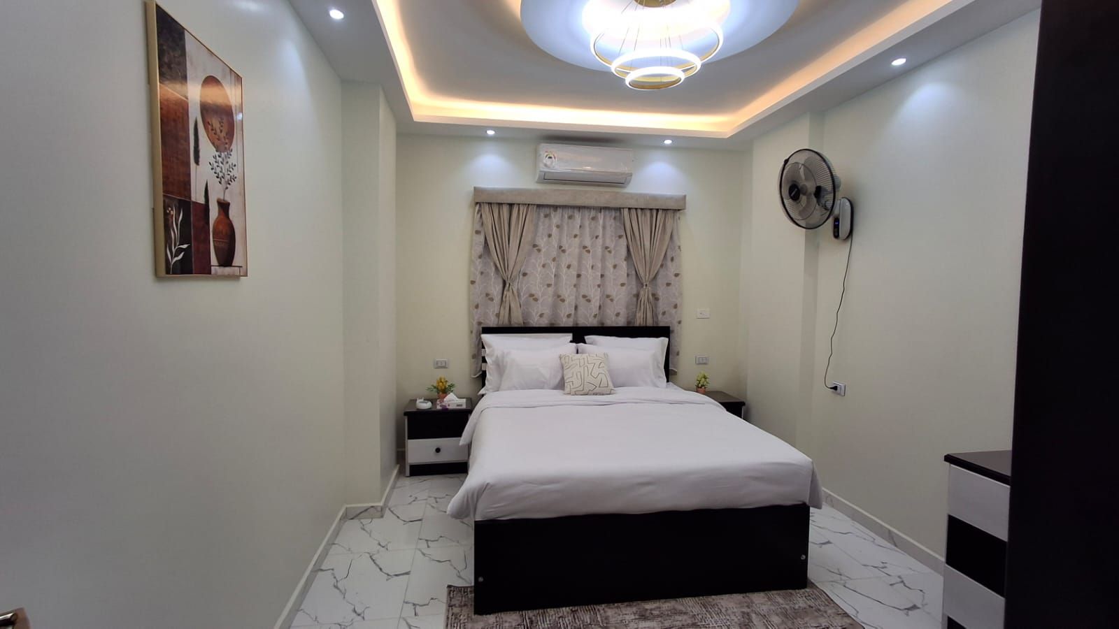 Een moderne slaapkamer met een bed, nachtkastjes, een airconditioner, een ventilator aan de muur en een plafondlamp.