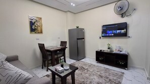 Een knus woongedeelte met een bank, een eettafel met stoelen, een koelkast, een koffiehoekje en een wandgemonteerde televisie.