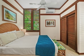 A hotel room with a bed, a ceiling fan, a window with blinds, a framed picture on the wall, and a door with a shutter. - Kiahuna 115 - Ground Floor - Corner Unit - Two ACs (Koloa)