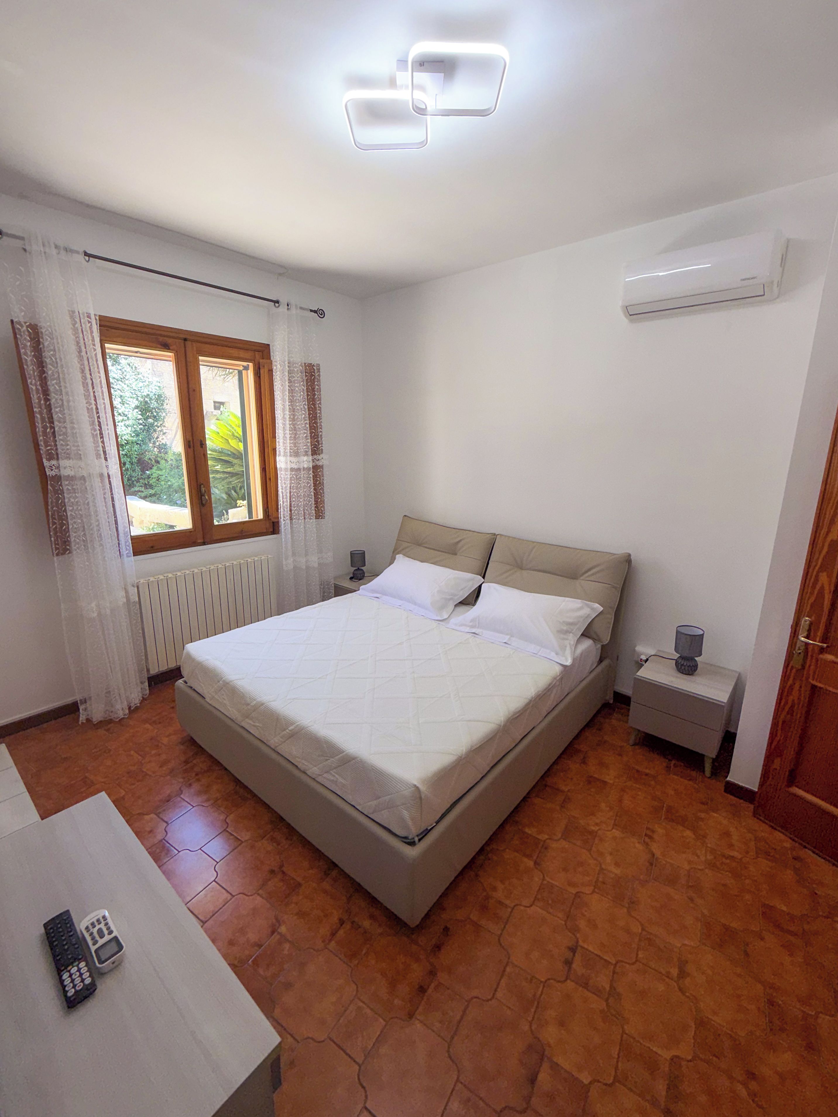 Une chambre à coucher comprenant un lit, une table de chevet, une lampe, une fenêtre avec des rideaux et un appareil de climatisation.