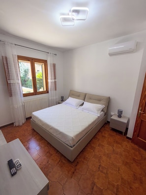 Ein Schlafzimmer mit einem Bett, einem Nachttisch, einer Lampe, einem Fenster mit Vorhängen und einer Klimaanlage.