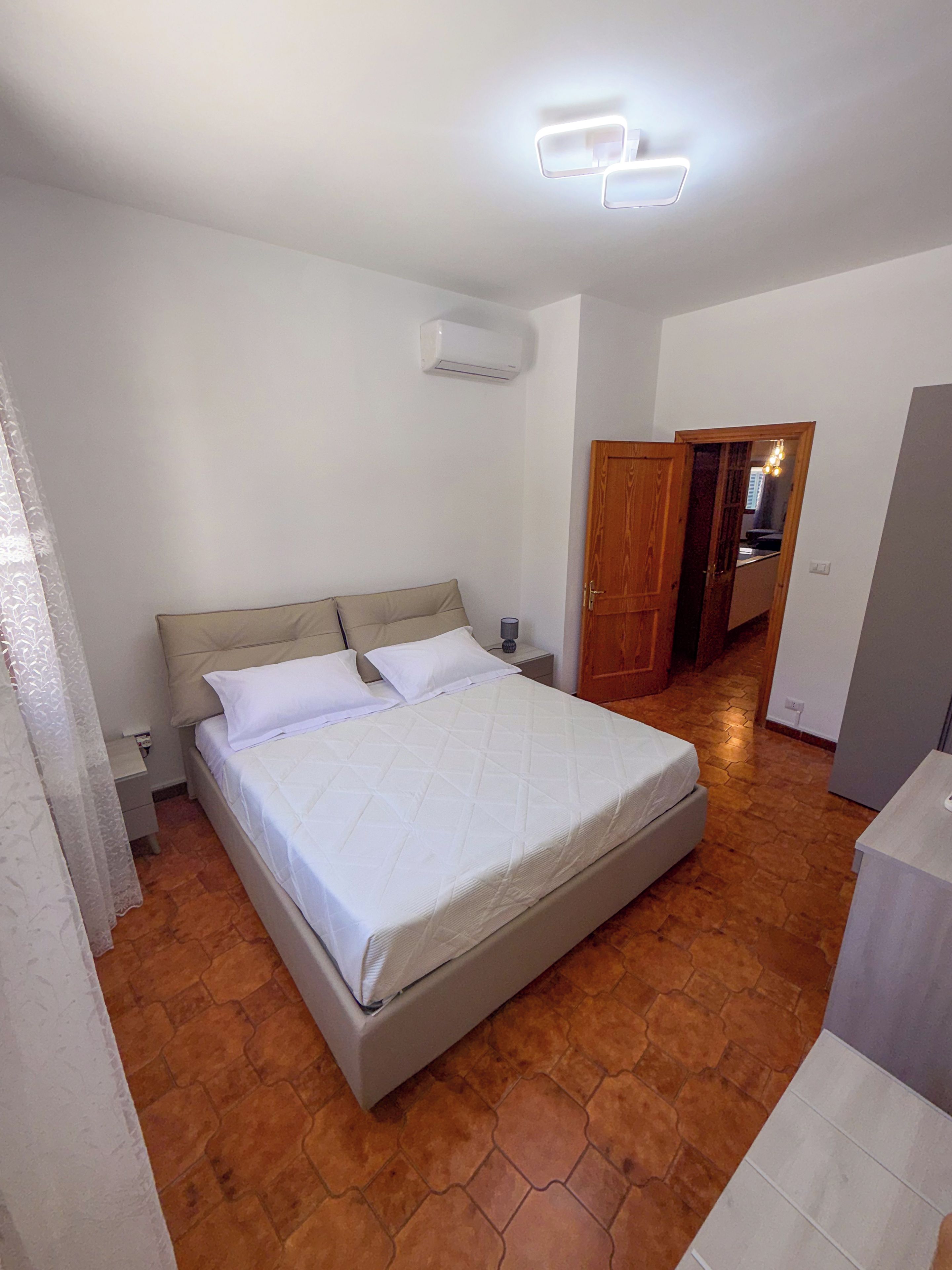 Een slaapkamer met een bed, een nachtkastje, een airconditioning en een houten deur naar de badkamer.