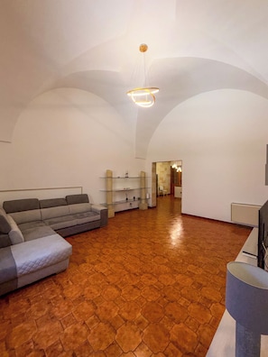 Un amplio salón con un sofá grande color gris, una mesa de centro y un mueble para el televisor. La habitación tiene piso de baldosas y un techo abovedado alto con una lámpara colgante.