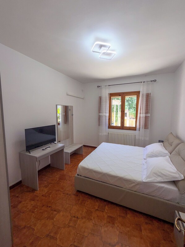 A bedroom with a bed, a TV, a desk, and a window with curtains.