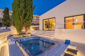 A modern outdoor hot tub area with a wooden deck, a large potted tree, and a building with large windows. - Villa Nostra w/ Pool, Gym & Tennis Court (Albufeira)