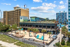 Un hotel frente a la playa con varios pisos, mesas al aire libre y una zona de piscina.