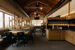 Een restaurantinterieur met houten meubels, een keukenhoek en grote ramen die natuurlijk licht binnenlaten.