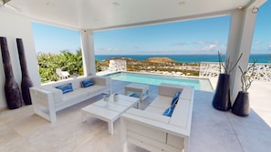 A modern outdoor lounge area with a pool, white furniture, and a view of the sea and landscape. - Vrbo Property (SAINT-MARTIN)