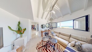A spacious living room with a high ceiling, a large window offering a view, a sofa, a coffee table, and a decorative rug. - Vrbo Property (SAINT-MARTIN)