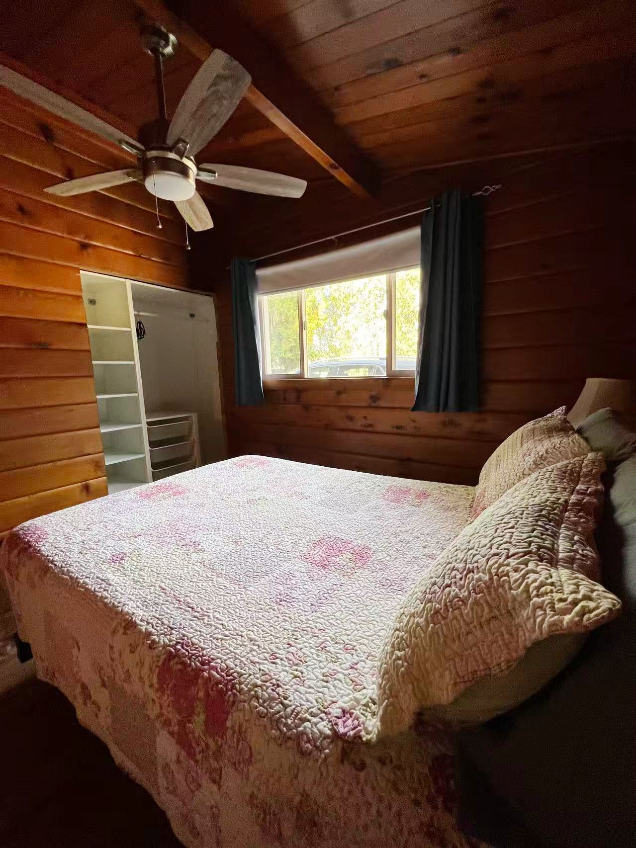 Un dormitorio con cama, ventilador de techo, ventana con cortinas y pared de madera.