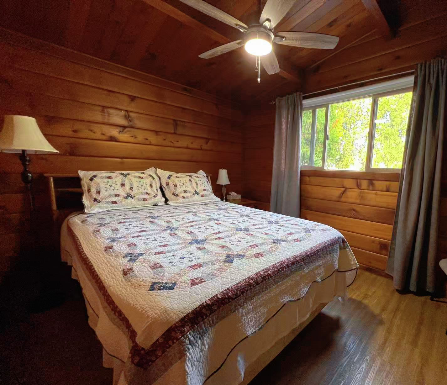 A wooden cabin bedroom with a bed, a ceiling fan, a window with curtains, and a lamp.