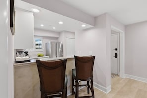 Una cocina moderna con refrigerador, un comedor con dos sillas de mimbre y una puerta que da a otra habitación.