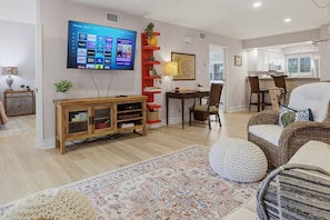 A modern living room with a flat-screen TV, a wooden entertainment unit, a dining area with a table and chairs, and a kitchen in the background. - Corner Condo Retreat | 1st Floor | Walk-Out Patio | Optional Saddlebrook Resort Amenities (Wesley Chapel)