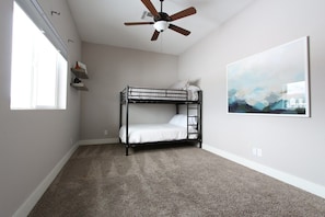 A room with a bunk bed, a ceiling fan, a window, and a painting on the wall.