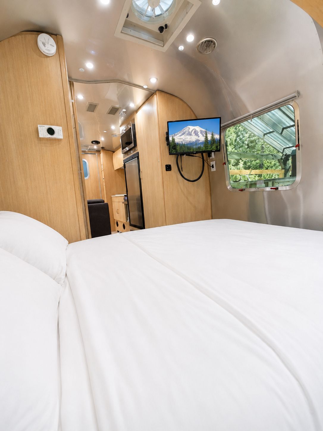 A compact, well-lit interior with a bed, a mounted TV, a window with a view of greenery, and a kitchenette area.