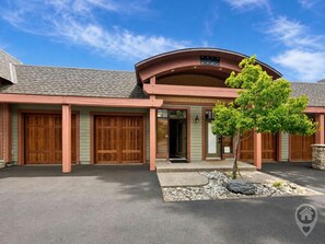 A two-story building with a curved roof and a central entrance. There are two garage doors on the ground floor and a small tree in front. - Ski-In/Ski-Out / Private Hot Tub & Mountain Views (Kimberley)