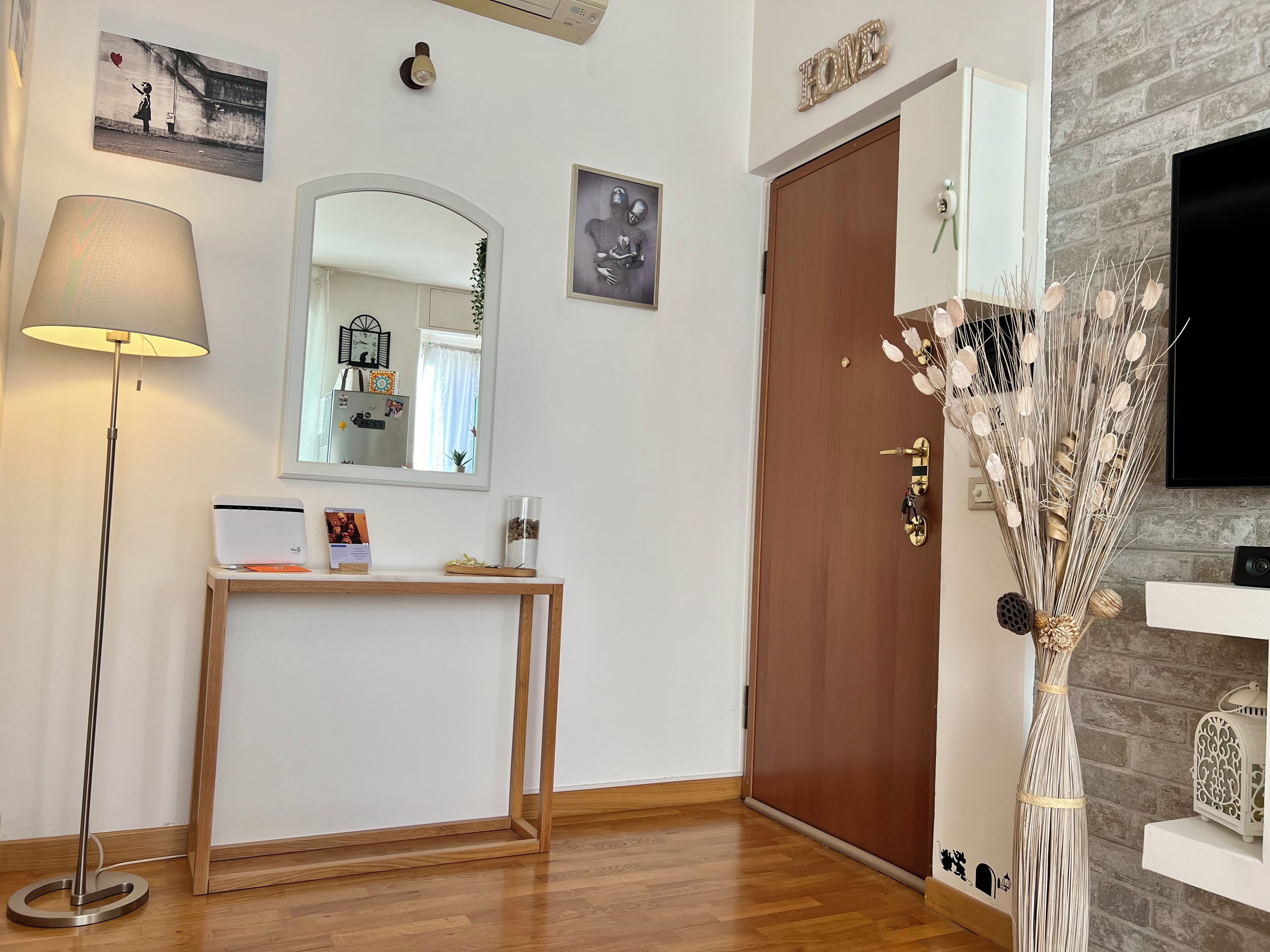 Une pièce avec un plancher en bois, un lampadaire, un miroir, une console sur laquelle sont posés divers objets, une porte et un téléviseur fixé au mur.