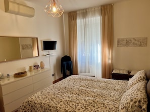 A bedroom with a bed, a dresser, a television, a window with curtains, and a wall-mounted air conditioner. - Nido Milano | La tua casa lontano da casa (Milano)