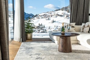 Una sala de estar con vistas a un paisaje nevado de montañas, un sofá, una mesa de centro con una botella y vasos, y una planta en maceta.