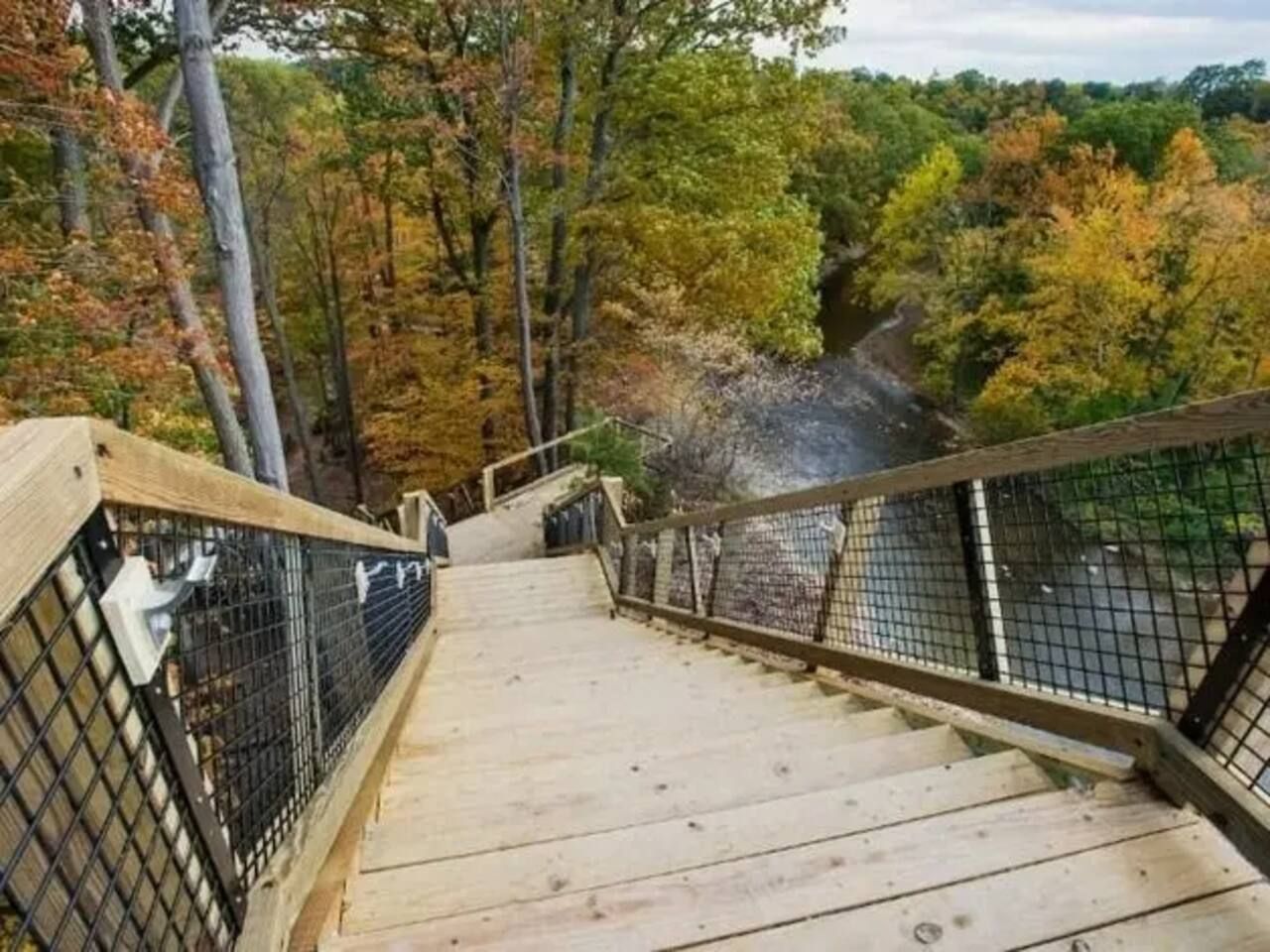 A wooden staircase with metal railings leading up a hill, surrounded by trees with autumn foliage.