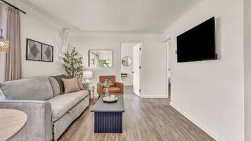 A modern living room with a grey sofa, a brown armchair, a coffee table, and a flat-screen TV mounted on the wall.