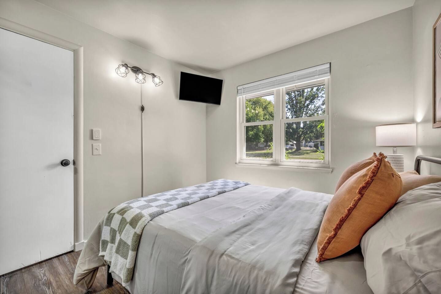 Un dormitorio con cama, ventana con persianas, televisor montado en la pared y una puerta.