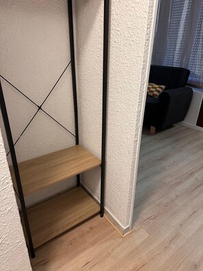 A corner of a room with a wooden shelf unit against a textured wall, a dark-colored sofa with a cushion, and a wooden floor.