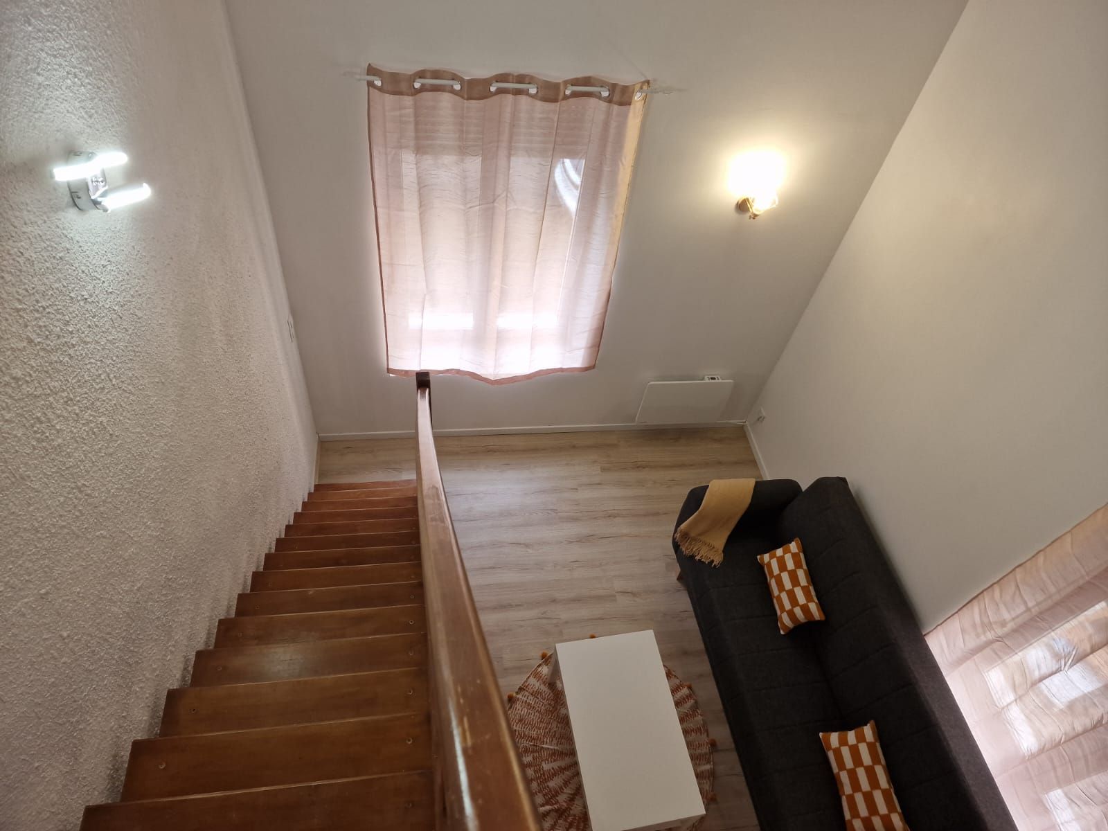 A staircase leading to a landing with a wooden floor, a white wall, a black sofa with patterned cushions, and a window with sheer curtains.