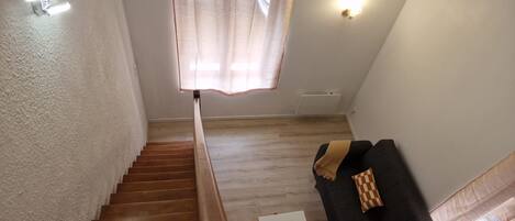 A staircase leading to a landing with a wooden floor, a white wall, a black sofa with patterned cushions, and a window with sheer curtains.